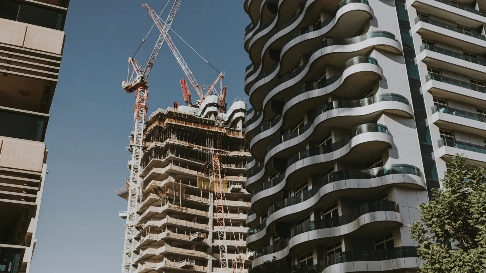 A residential tower under construction with cranes beside a finished apartment building with curved balconies in Dubai