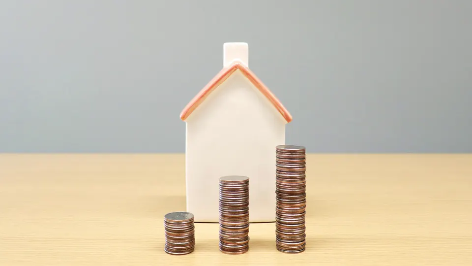 A small white model house beside three stacks of coins growing in height on a wooden table