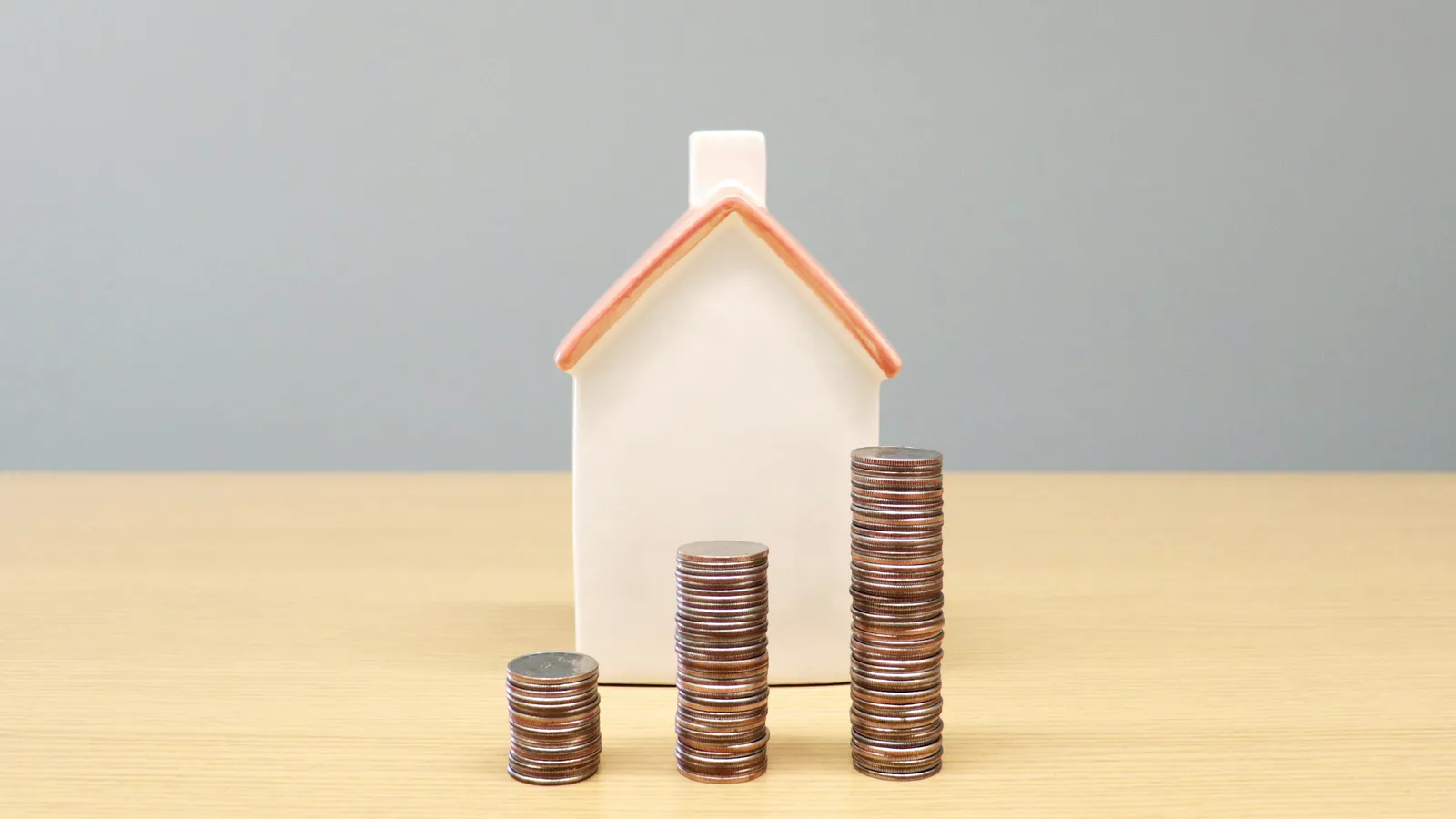 A small white model house beside three stacks of coins growing in height on a wooden table